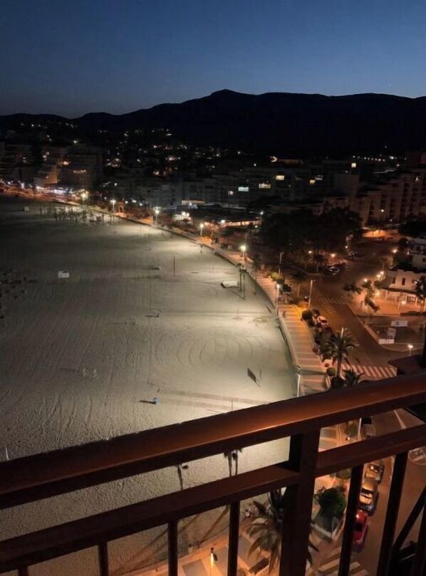 A nighttime view of a beach with a clear sky, city lights in the distance, and a mountain range in the background.