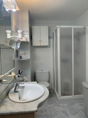 Un baño con lavamanos blanco, espejo, inodoro y ducha con mampara de vidrio.