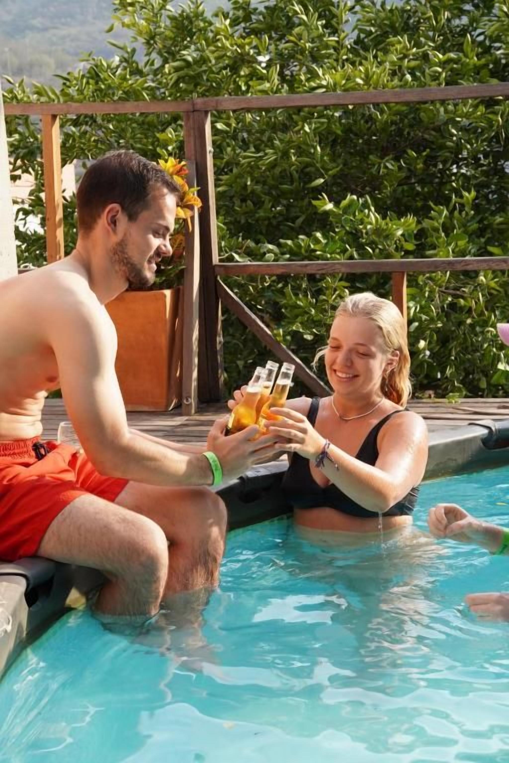 Dos personas en una piscina con cubierta de madera y follaje al fondo.