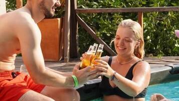 Two people in a swimming pool with a wooden deck and greenery in the background.
