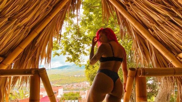 A person standing in a thatched-roof structure with a view of a swimming pool and landscape. - Traveler Hostel (San Gil)