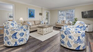 A living room with a beige sofa, two ottomans with a nautical theme, a coffee table, and a TV mounted on the wall.
