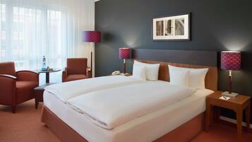 A hotel room with a large bed, two bedside tables, a chair, a small table with a lamp, and a window with sheer curtains.
