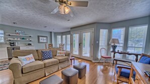 A spacious living room with a beige sofa, ottomans, a dining area with a table and chairs, and large windows allowing natural light.