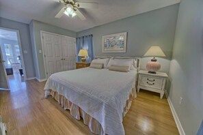 A bedroom with a large bed, a nightstand with a lamp, a ceiling fan, and a door leading to another room. - A Block Away Bliss: Steps from Seaside & Sand! (Santa Rosa Beach)