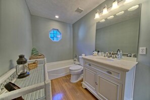 Une salle de bain avec une baignoire, des toilettes et un meuble-lavabo avec un miroir. Il y a une fenêtre à motifs hexagonaux et un panier en osier posé sur le sol.