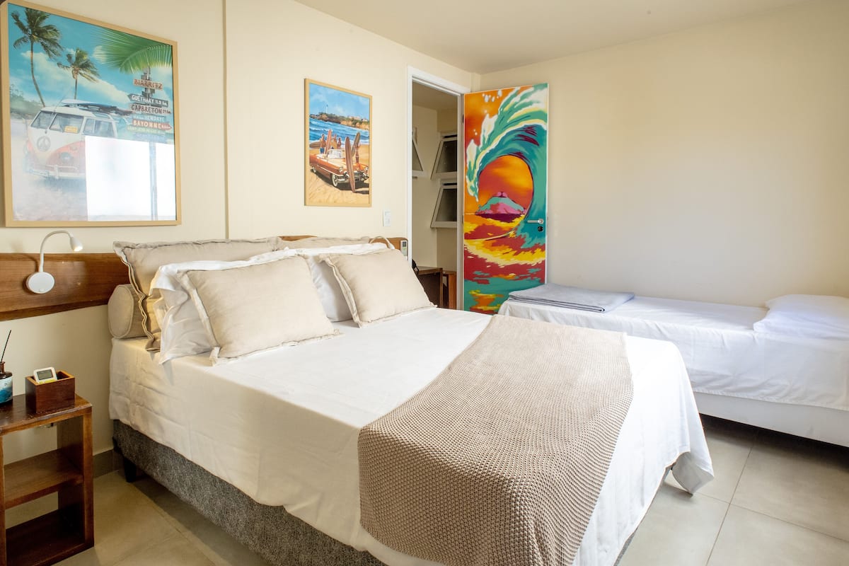 Quarto de hotel com uma cama grande, uma cama menor, uma mesa de cabeceira e uma parede com duas pinturas.