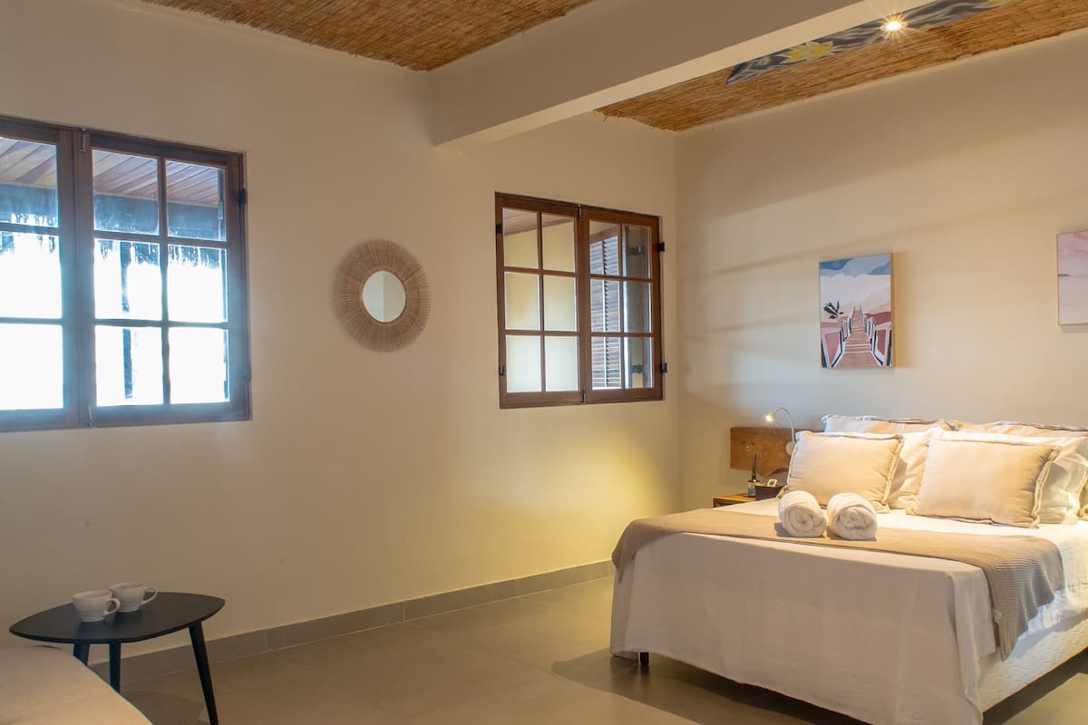 Quarto com cama, mesa de cabeceira, espelho redondo e janela.
