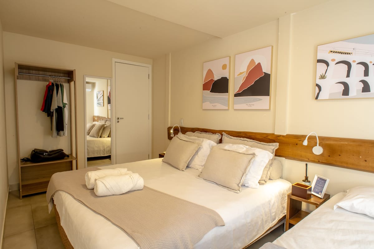 Quarto de hotel com uma cama grande, cabeceira de madeira, mesa de cabeceira com abajur, aparador, guarda-roupa e duas obras de arte emolduradas na parede.