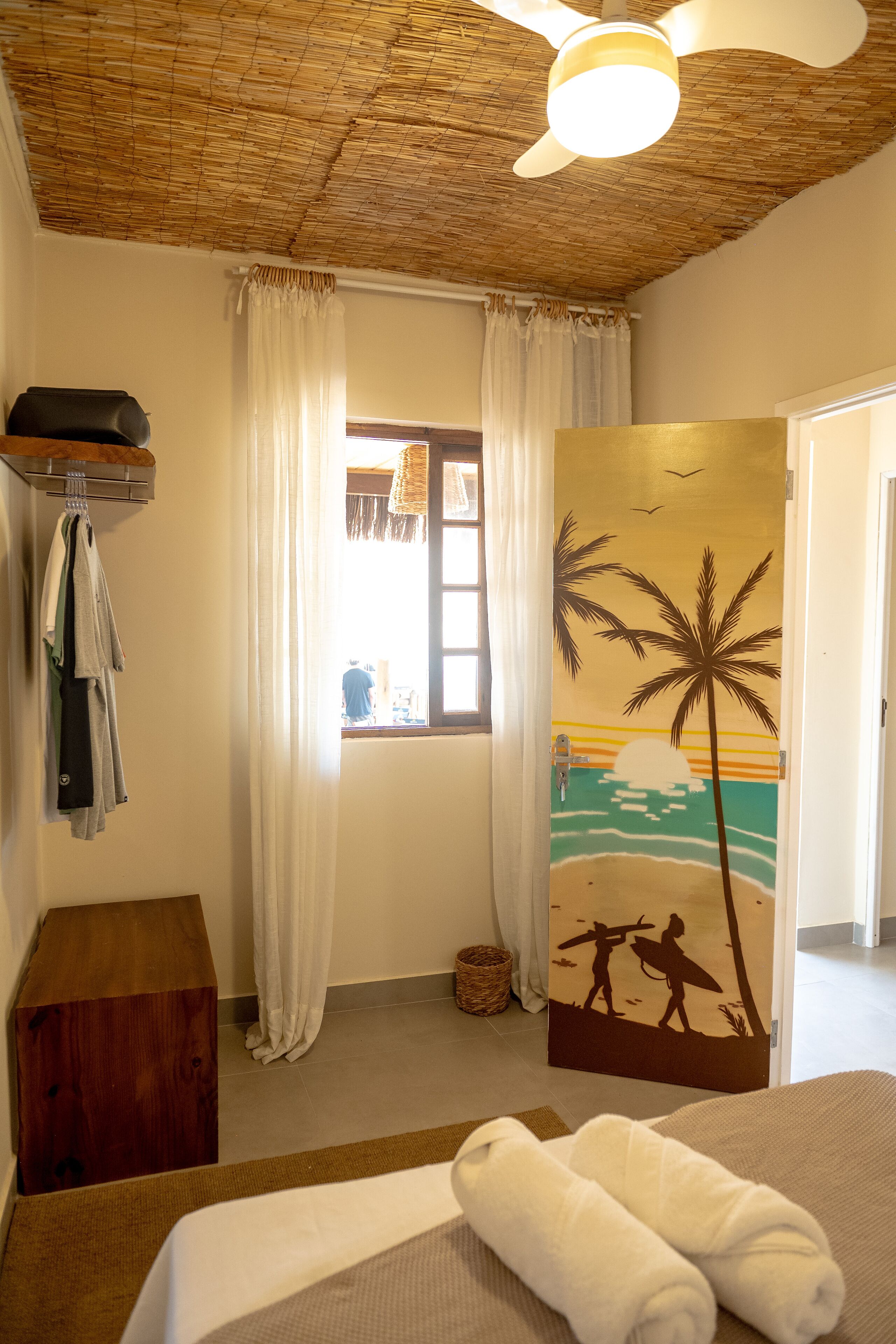 Une chambre avec un lit, un ventilateur de plafond, une porte décorée sur le thème de la plage et une fenêtre avec des rideaux.