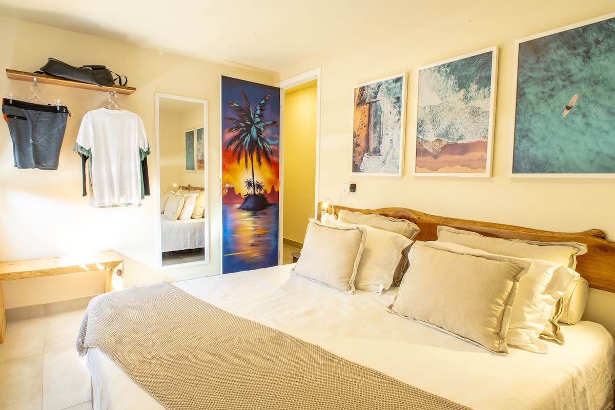 Quarto de hotel com uma cama grande, cabeceira de madeira e uma pintura do pôr do sol. Há um banco e uma prateleira com malas e roupas.