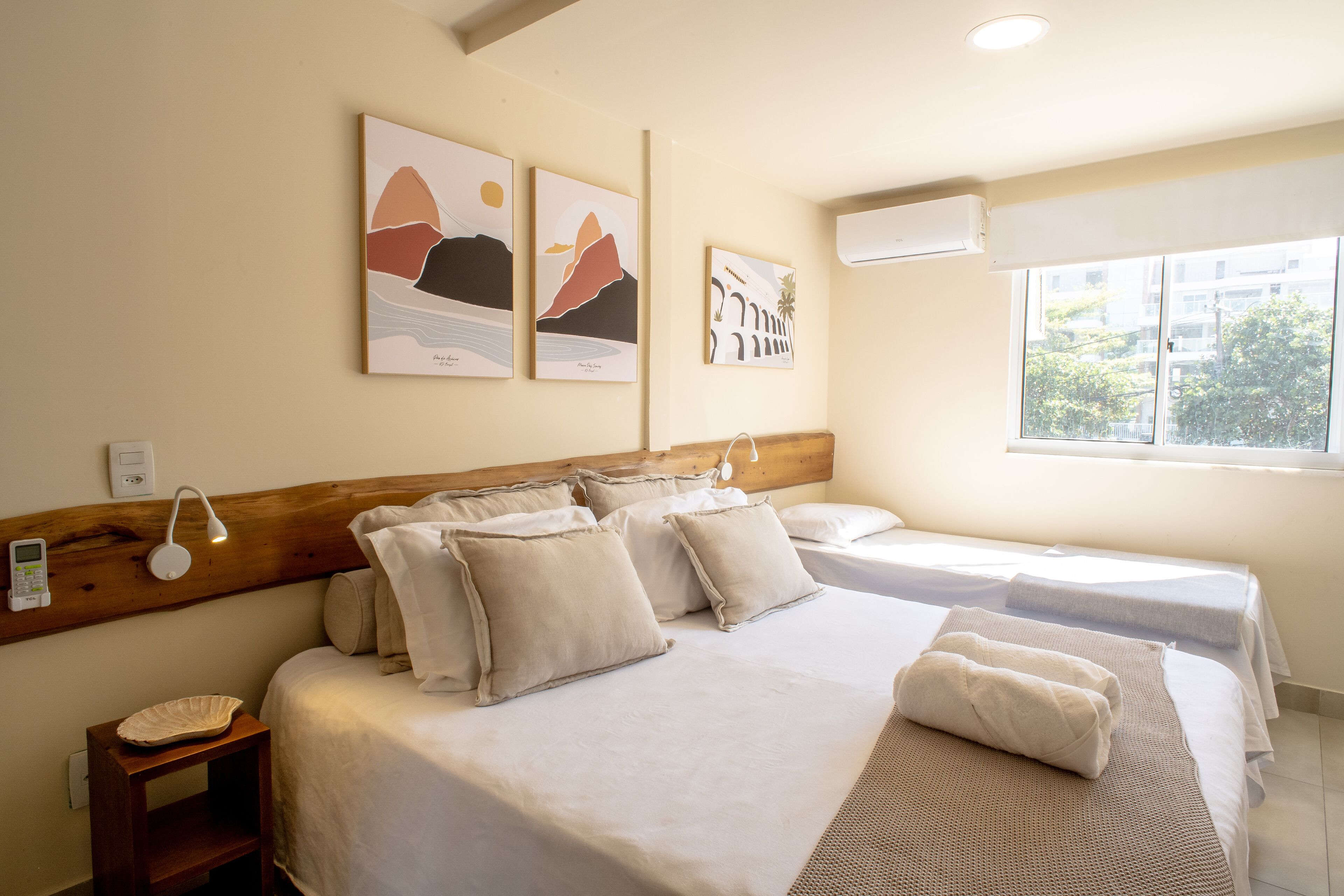 A hotel room with two beds, a bedside table, a lamp, artwork on the wall, and a window with a view.