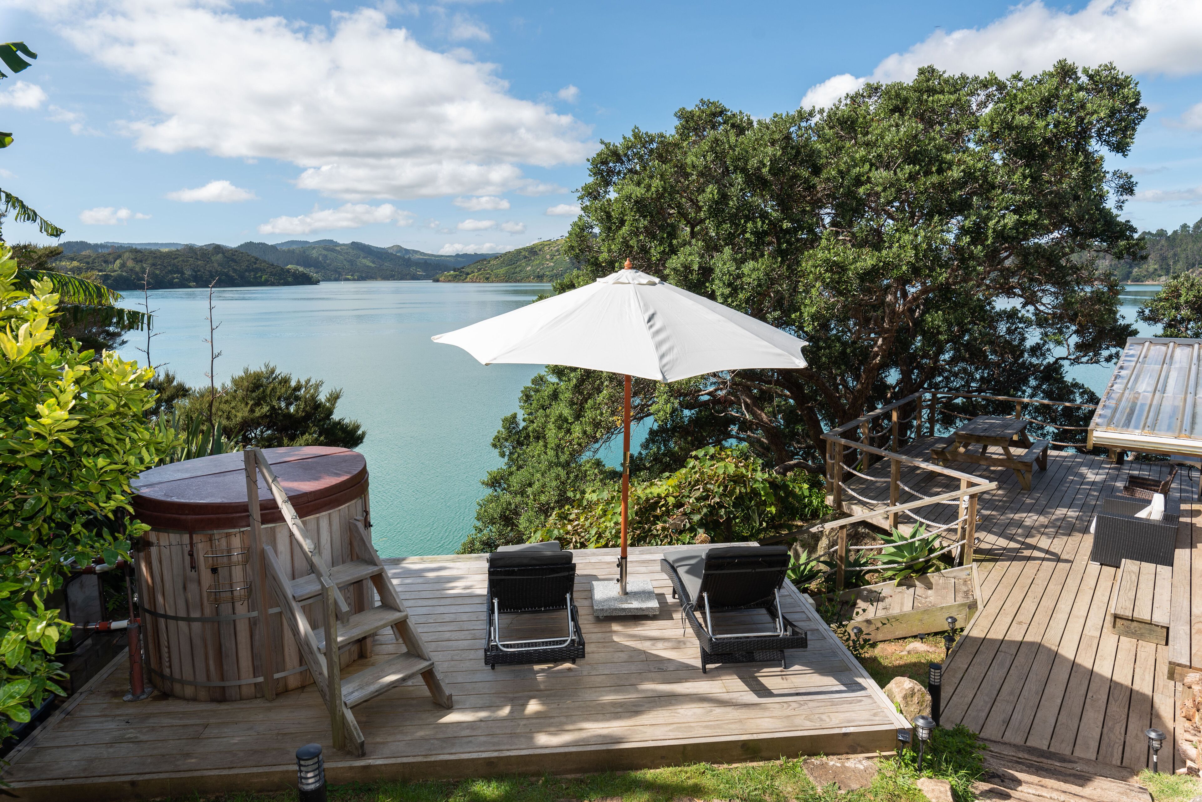 A wooden deck with two lounge chairs and a white umbrella, overlooking a lake with mountains in the background.