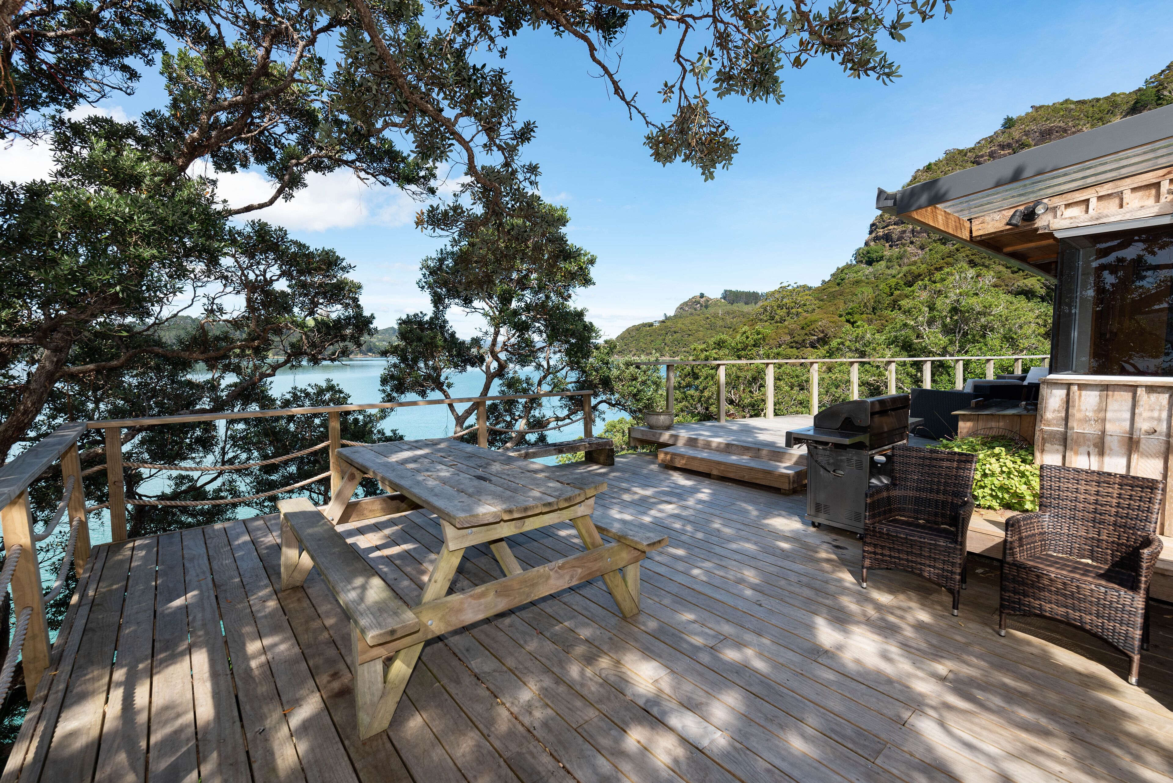 Un terrazzo in legno con tavoli da picnic e sedie in vimini, affacciato su uno specchio d'acqua e delle colline.