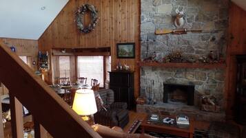 A cozy living room with a stone fireplace, wooden walls, and a dining area with a table and chairs.