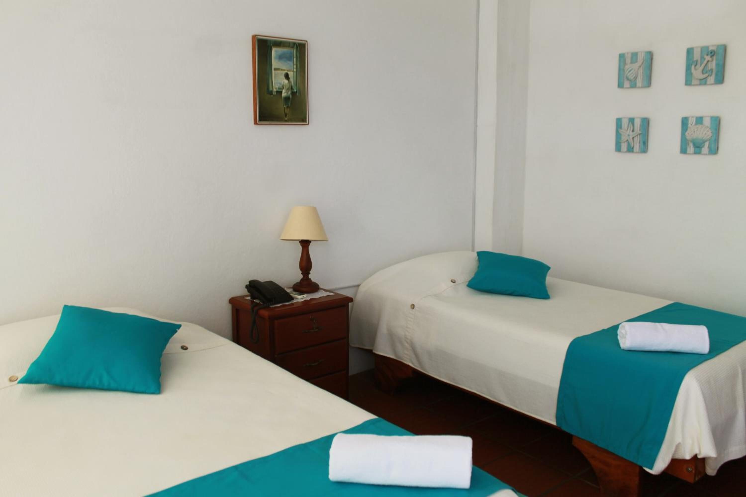 A hotel room with two single beds, each with a teal pillow and blanket, a wooden nightstand with a lamp, and a small painting on the wall.
