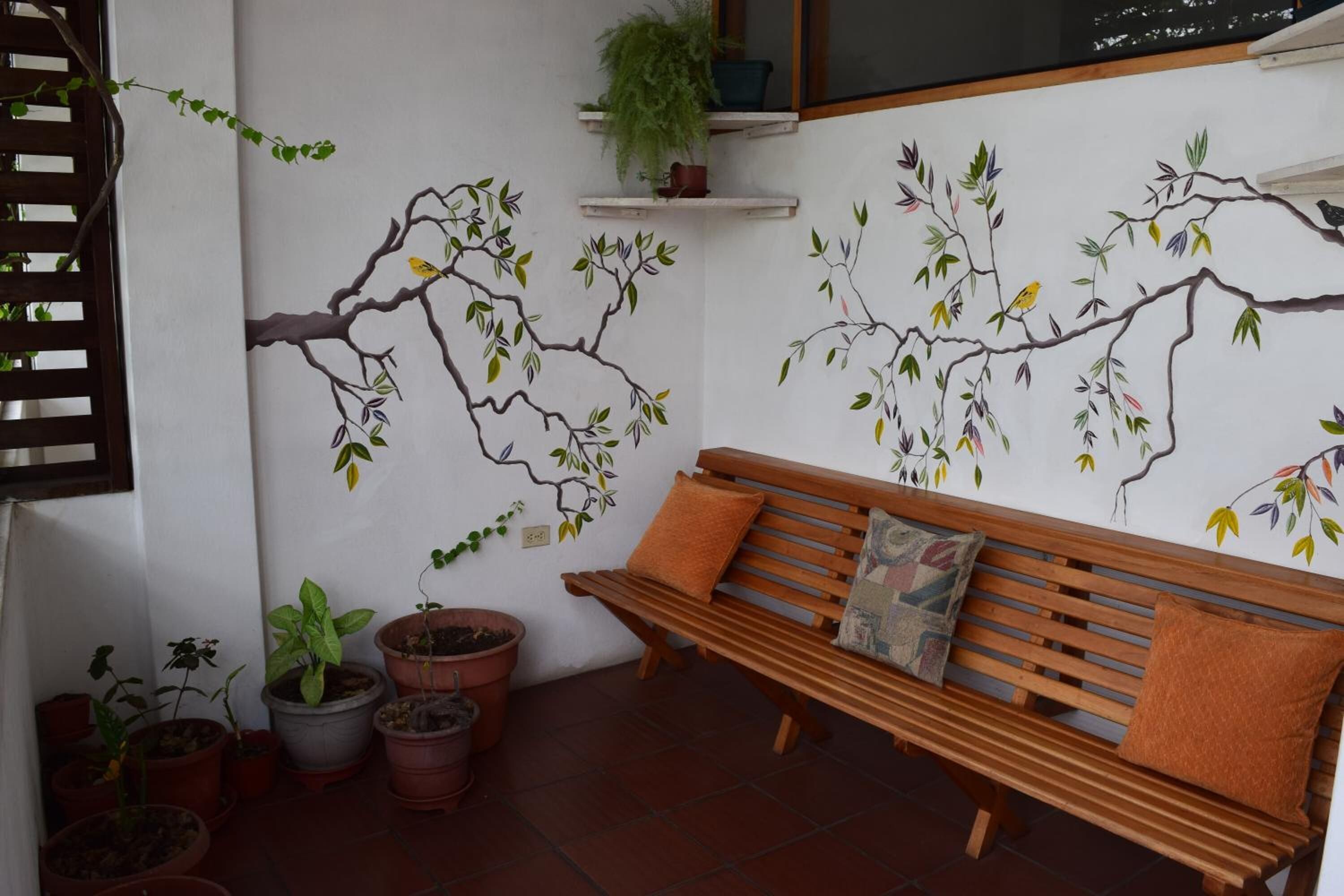 Un banco de madera con cojines, plantas en macetas y un mural en la pared con ramas y hojas.