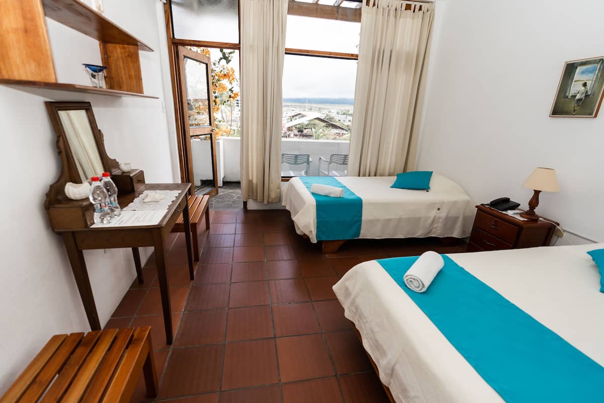 Quarto de hotel com duas camas, um banquinho de madeira, uma mesa com um espelho e vista da cidade através de amplas janelas.