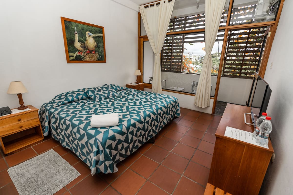 Quarto com colcha estampada, mesa de cabeceira com abajur, escrivaninha de madeira com garrafa de água e janela com vista para a cidade.