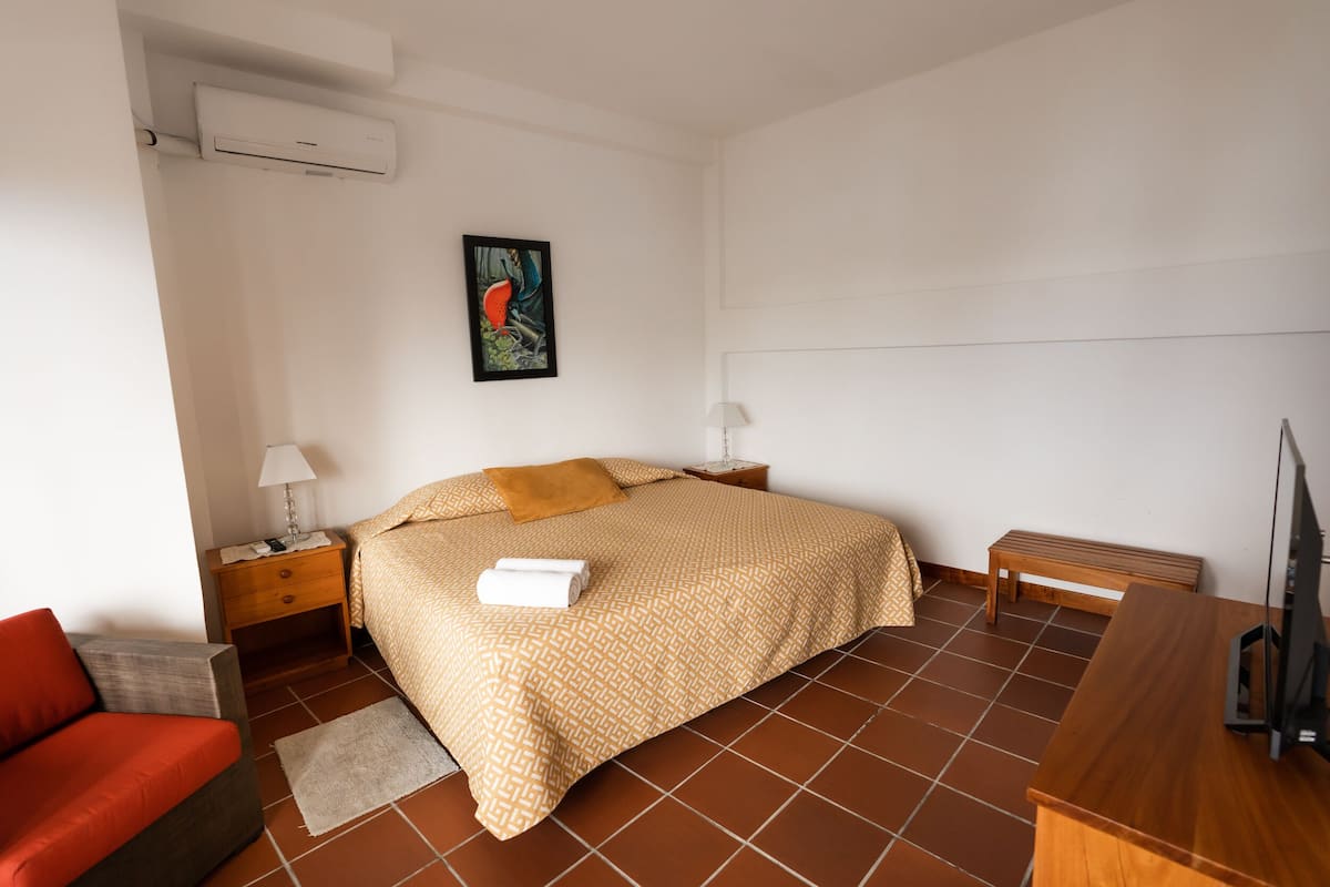 Um quarto com cama, mesa de cabeceira, luminária, banco e televisão.