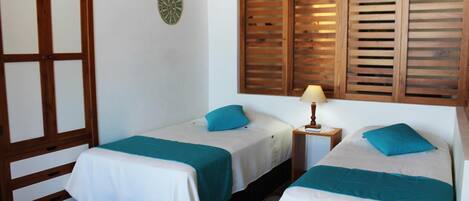Une chambre avec deux lits simples, chacun agrémenté d’un oreiller turquoise et de linge de lit blanc. On y trouve une armoire en bois avec une porte en verre, une petite table avec une lampe et une horloge murale.