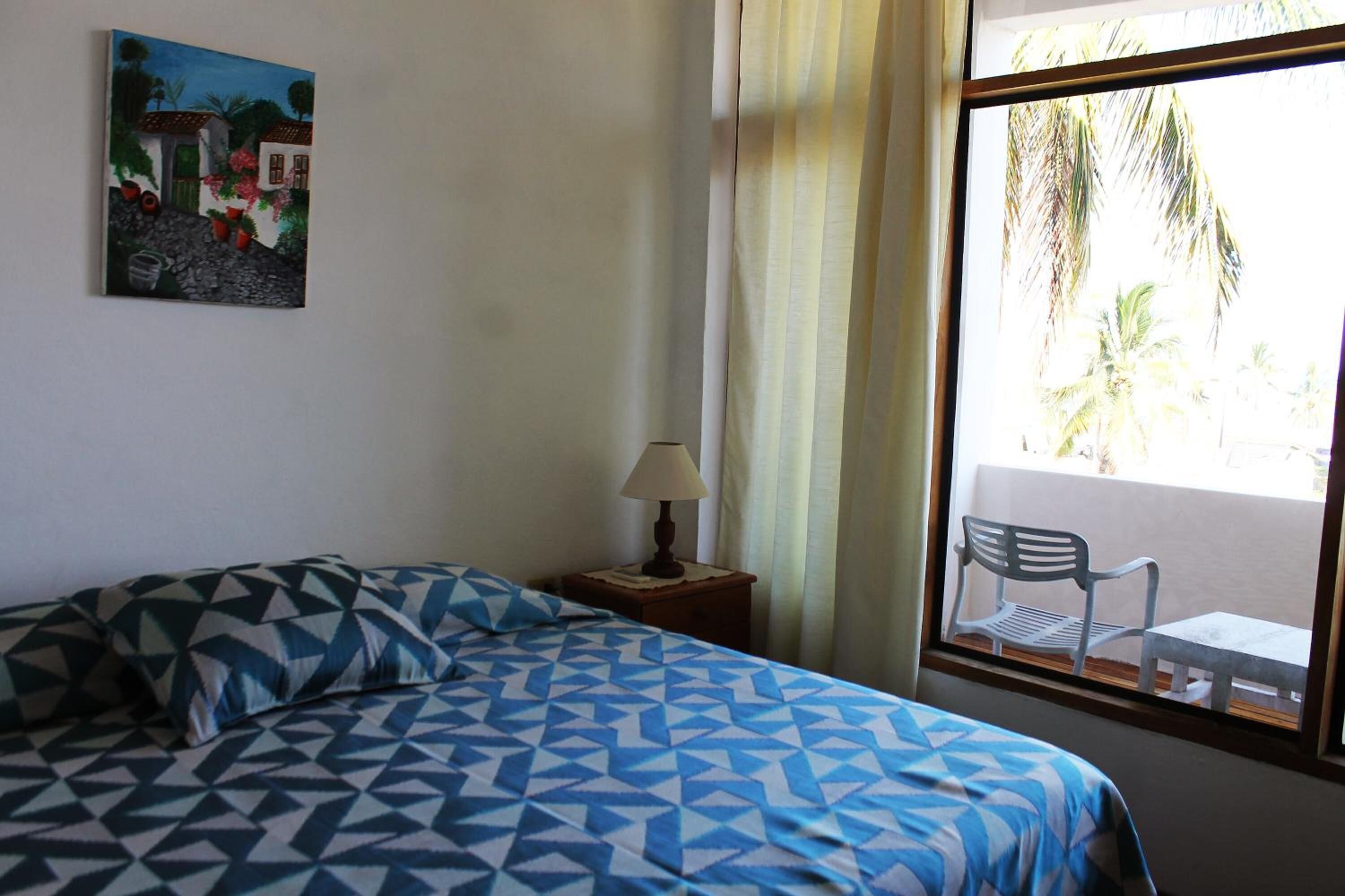 A bedroom with a bed, a nightstand with a lamp, a window with a view of palm trees, and a painting on the wall.