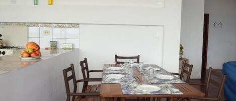 A dining area with a table set for four, surrounded by wooden chairs. A fruit bowl with apples and a pumpkin is on the counter.