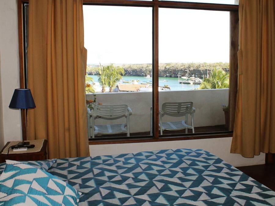 A room with a bed, a window with curtains, a lamp, and a view of a beach and boats.