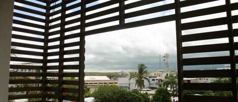 Una habitación con vistas a través de una ventana con persianas horizontales, mostrando un paisaje urbano con edificios y palmeras.