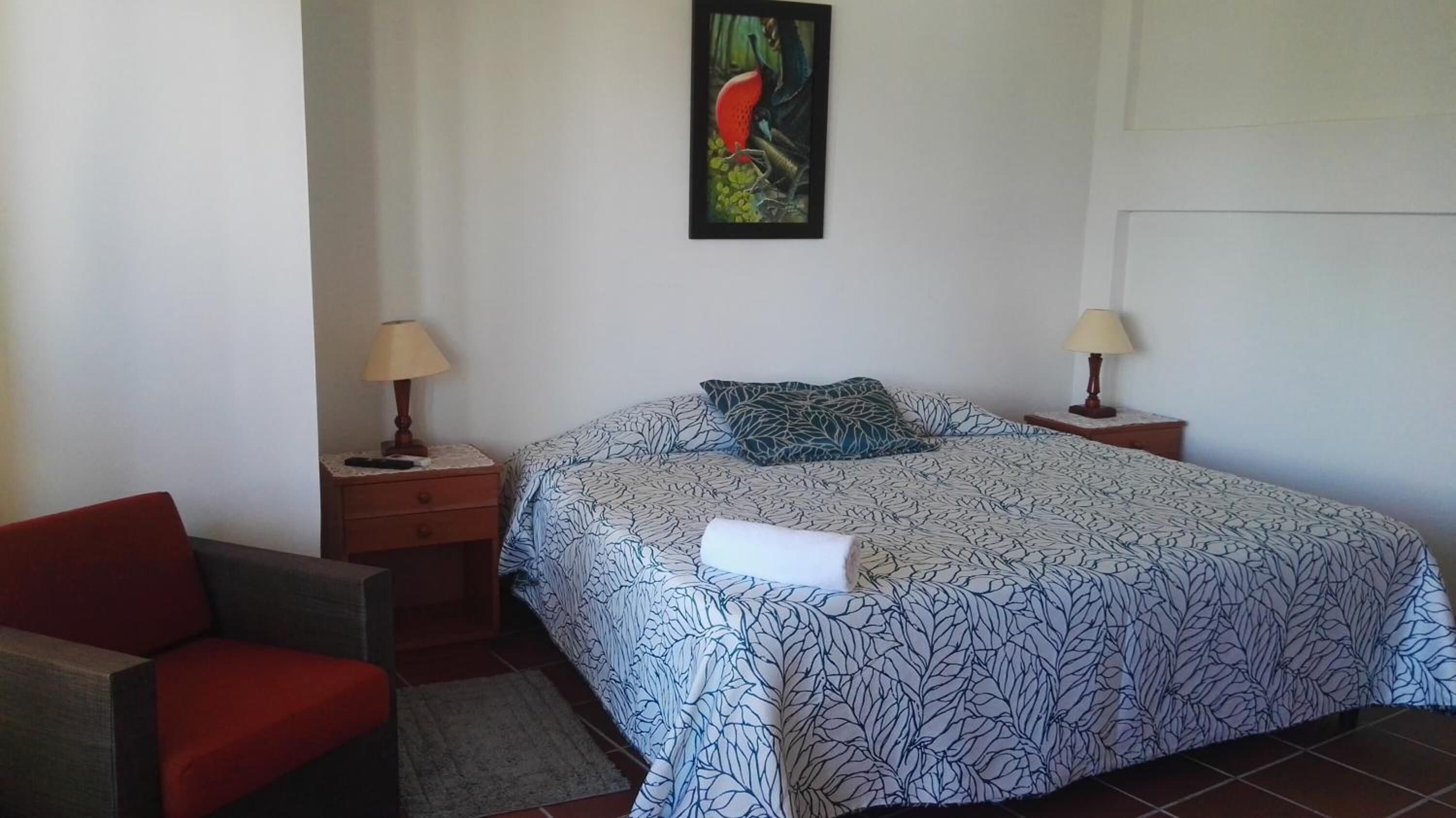 Un dormitorio con una cama, dos mesitas de noche, una silla y un cuadro en la pared.