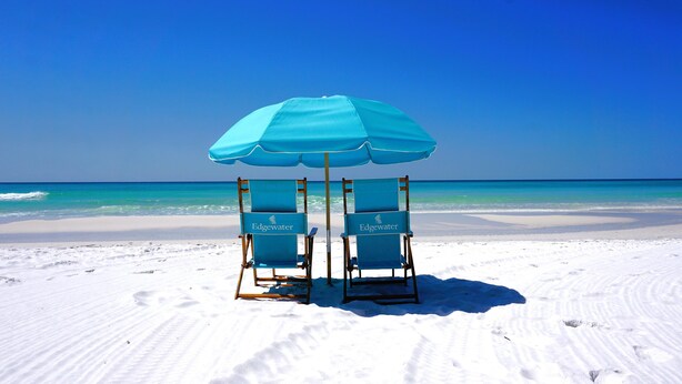 Twee strandstoelen onder een blauwe parasol op een zandstrand met helderblauw water en lucht.