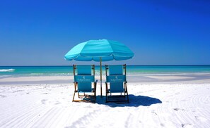 Two beach chairs under a blue umbrella on a sandy beach with clear blue water and sky.