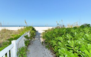 Een zandpad dat naar een strand leidt, met een witte omheining aan één kant en weelderige groene beplanting aan de andere kant.