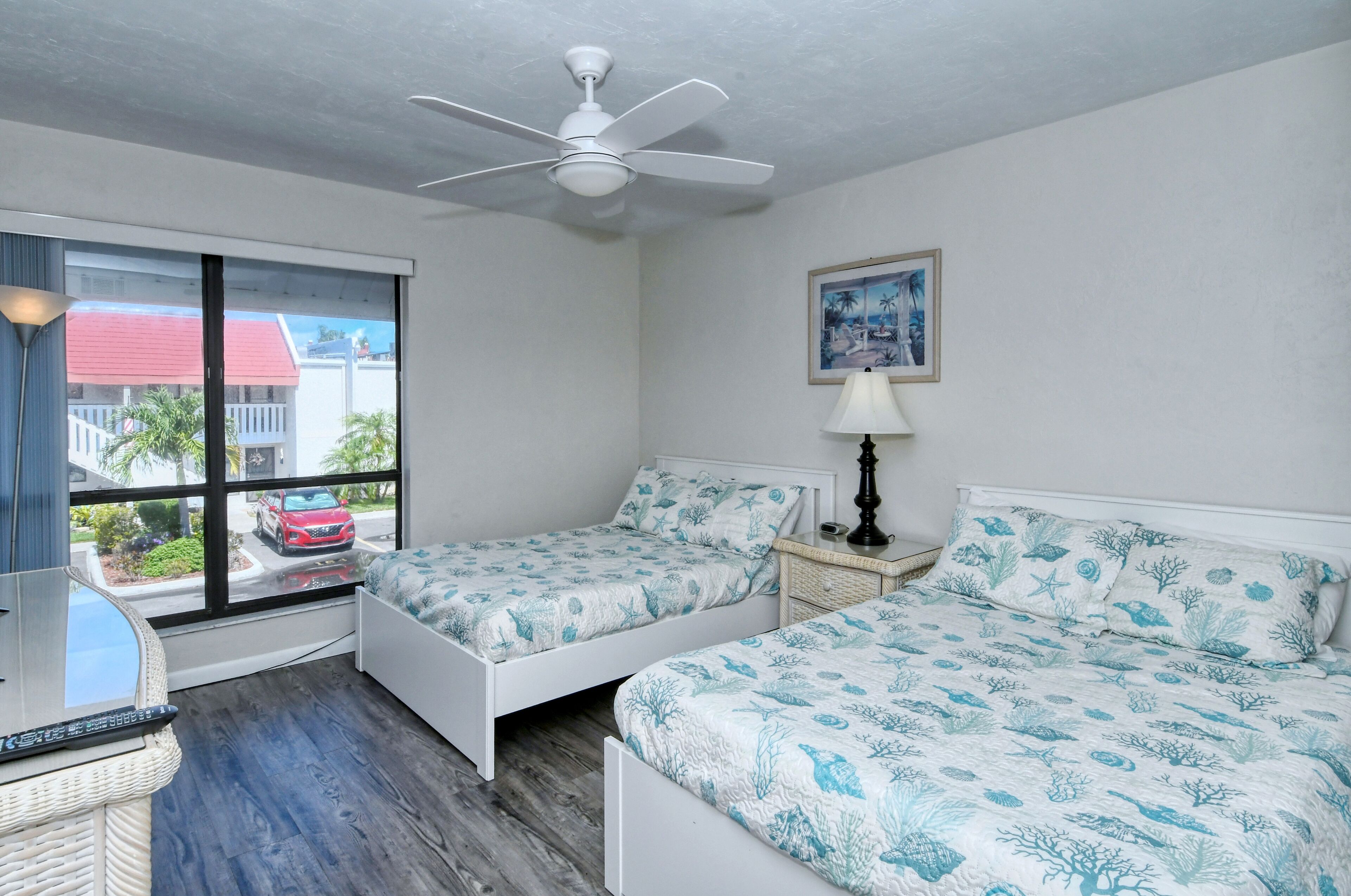 A bedroom with two beds, a ceiling fan, a window with a view of a red car and buildings, and a lamp on a nightstand.
