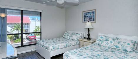 A bedroom with two beds, a ceiling fan, a window with a view of a red car and buildings, and a lamp on a nightstand.