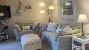 Ein Wohnzimmer mit grauem Sofagarnitur, einem Couchtisch, einem Fernseher, einem Bücherregal und einem gerahmten Bild an der Wand.