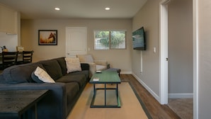 A living room with a sofa, a coffee table, a TV mounted on the wall, and a window with a view of greenery.