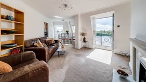 A spacious living room with a brown leather sofa, a coffee table, and a fireplace. There is a balcony with a railing and a view of the outdoors. - River View, London Zone 2 Hammersmith, 2 Bedroom / 2 Bath (London)