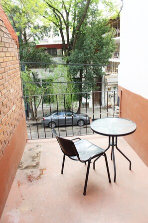 A balcony with a metal table and chair, a brick wall, and a view of a street with a car and trees.