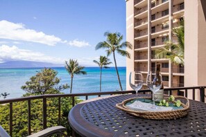 A balcony with a view of the ocean, a table with two wine glasses, and a building with multiple balconies.