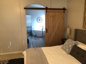 A bedroom with a bed, a bedside lamp, a patterned pillow, a wooden door, and a view into a bathroom with a bathtub and a shower.
