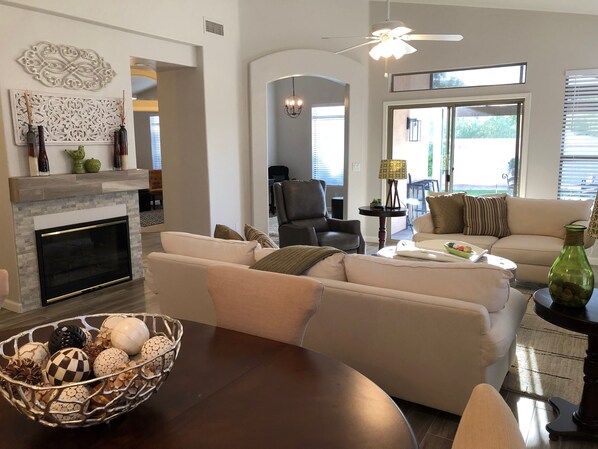 A living room with a fireplace, a sofa set, a dining table with a decorative bowl, and a ceiling fan.