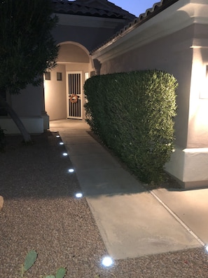 A pathway leading to a house entrance with a wreath on the door, a hedge on the right, and a tree on the left.