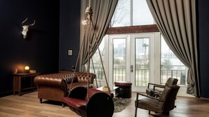 A room with a hanging swing chair, a leather sofa, and a chair with a metal frame. There is a large window with curtains, a wooden floor, and a wall-mounted light fixture. - Huge Sunlit Loft w/ Indoor Swing and Skyline Views (Houston)