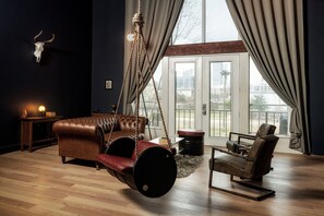 A room with a hanging swing chair, a leather sofa, and a chair with a metal frame. There is a large window with curtains, a wooden floor, and a wall-mounted light fixture.