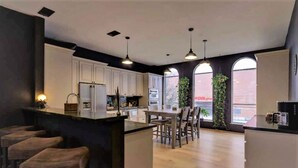 A modern kitchen with white cabinets, a central island with stools, pendant lighting, and large windows with plants. - Huge Sunlit Loft w/ Indoor Swing and Skyline Views (Houston)