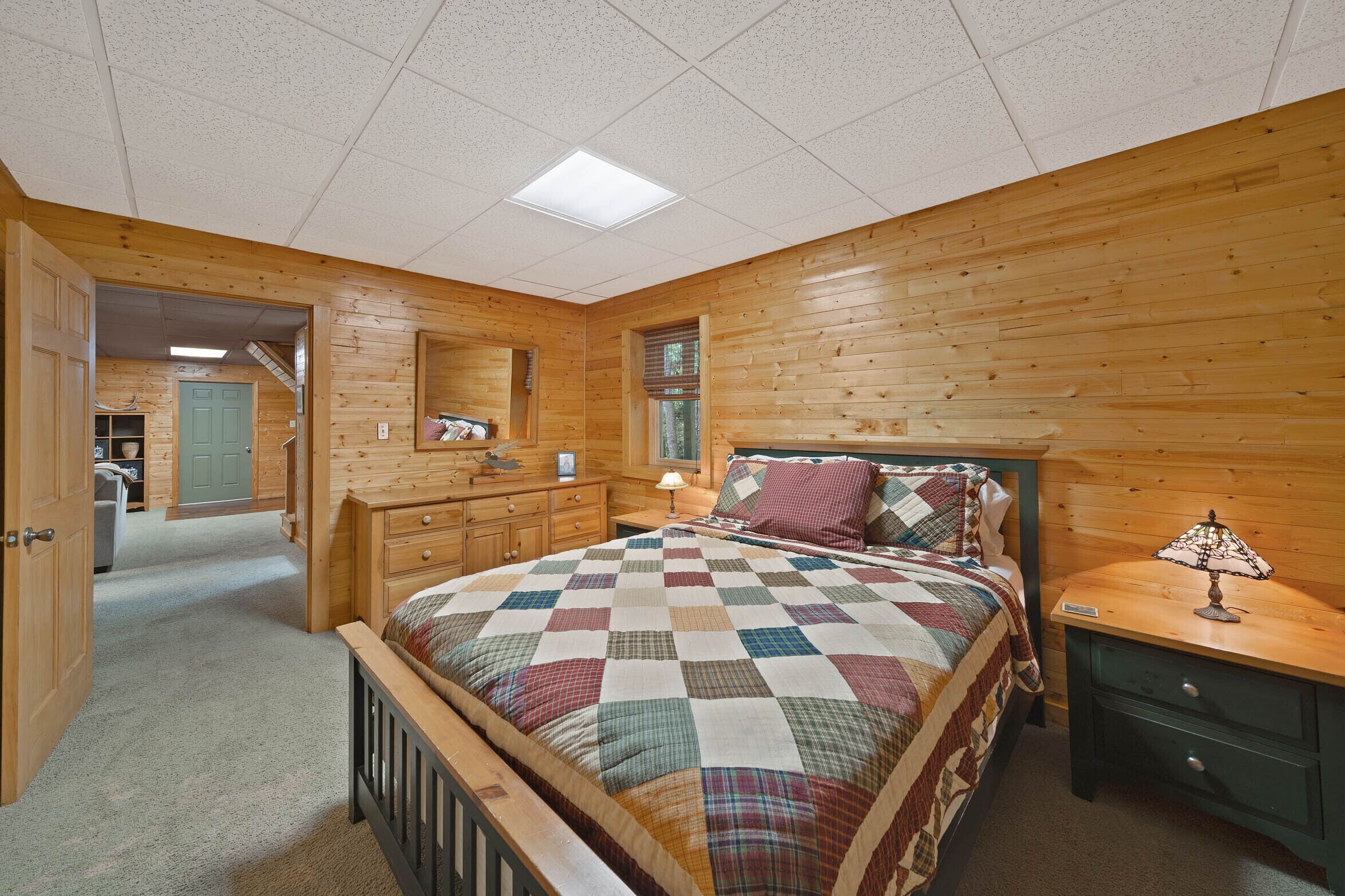 A bedroom with a wooden floor, a bed with a checkered quilt, a nightstand, a dresser, and a door leading to another room.