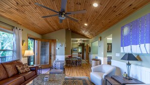 A cozy living room with a brown leather sofa, a white armchair, and a coffee table. A ceiling fan is visible, and the room has wooden flooring and a large window. - Chipmunk View Lodge - Amazing views, hot tub, and manmade waterfall! (Blowing Rock)
