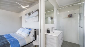 A modern bedroom with a bed, a bedside table, a nightstand, a ceiling fan, and a bathroom with a shower, sink, and mirror.
