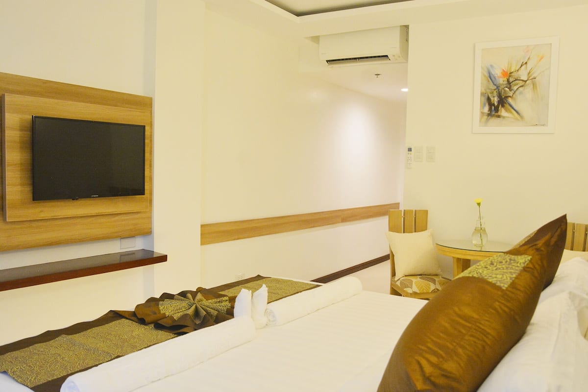 A hotel room with a bed, a television mounted on the wall, a small table with a vase, and a painting on the wall.